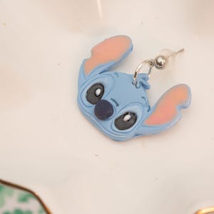 Pu&ograve; includere: Un singolo orecchino blu di Stitch con orecchie interne rosa e un naso blu scuro. L'orecchino &egrave; un design di personaggi dei cartoni animati con un perno e un anello color argento. L'orecchino &egrave; appoggiato su un piatto bianco smerlato con bordo dorato.