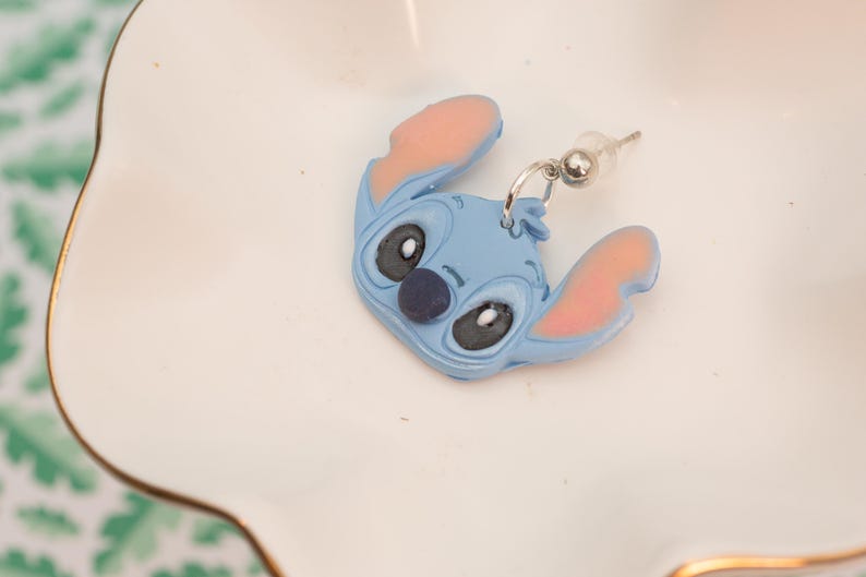 Pu&ograve; includere: Un singolo orecchino blu di Stitch con orecchie interne rosa e un naso blu scuro. L'orecchino &egrave; un design di personaggi dei cartoni animati con un perno e un anello color argento. L'orecchino &egrave; appoggiato su un piatto bianco smerlato con bordo dorato.