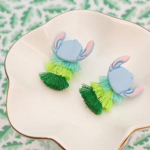 May include: Handmade earrings with a light blue character design and pink ears, paired with layered green and blue tassels. The earrings are presented on a white, scalloped dish with a gold trim.