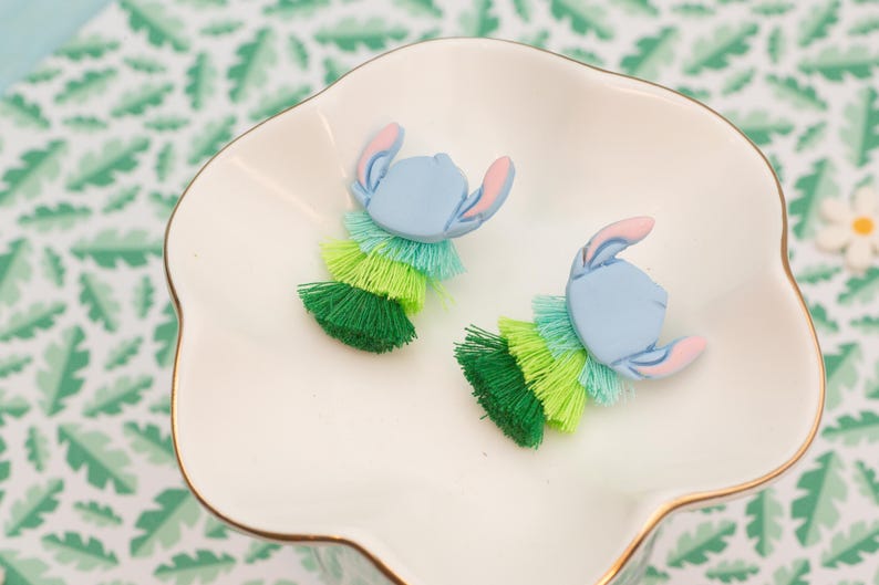 May include: Handmade earrings with a light blue character design and pink ears, paired with layered green and blue tassels. The earrings are presented on a white, scalloped dish with a gold trim.