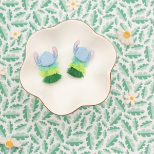 May include: Two blue and green Stitch-themed earrings with a layered fringe design, presented on a white, scalloped dish. The earrings feature blue heads with pink ears and are surrounded by green and yellow fringe. The background is a green and white leaf pattern.