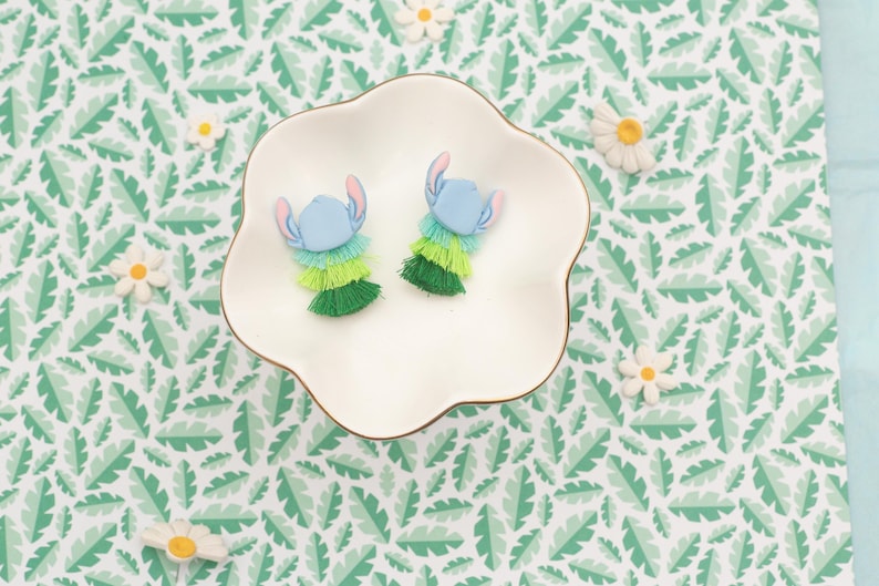 May include: Two blue and green Stitch-themed earrings with a layered fringe design, presented on a white, scalloped dish. The earrings feature blue heads with pink ears and are surrounded by green and yellow fringe. The background is a green and white leaf pattern.