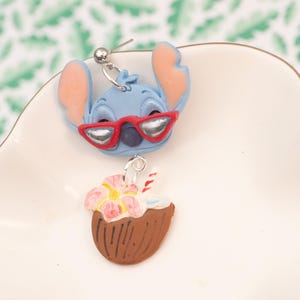 May include: A pair of novelty earrings featuring a blue cartoon character with red sunglasses and a coconut drink. The earrings are set on a white surface with a green and white leaf pattern in the background.