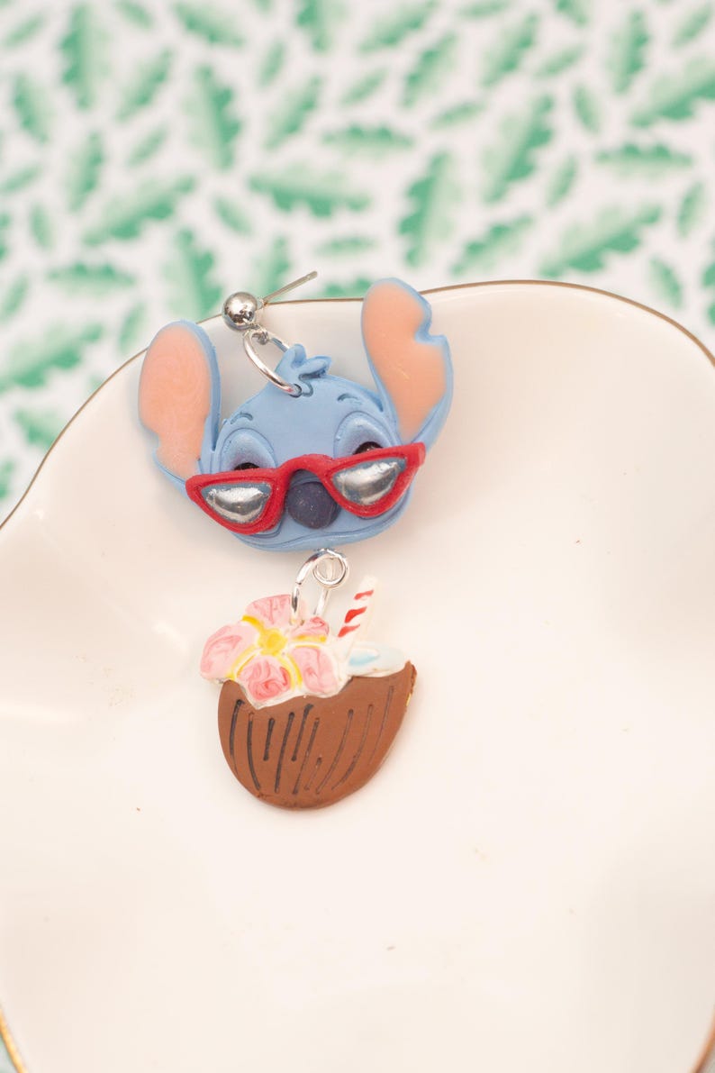 May include: A pair of novelty earrings featuring a blue cartoon character with red sunglasses and a coconut drink. The earrings are set on a white surface with a green and white leaf pattern in the background.