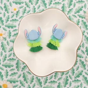 May include: A pair of blue and green Stitch-themed earrings with layered fringe. The earrings are displayed on a white, scalloped dish with a gold rim, set against a tropical leaf pattern background. The earrings feature blue heads and pink ears.