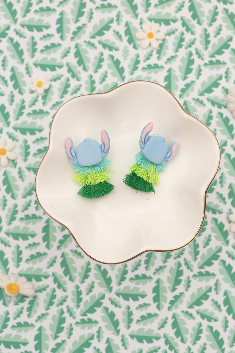 May include: A pair of blue and green Stitch-themed earrings with layered fringe. The earrings are displayed on a white, scalloped dish with a gold rim, set against a tropical leaf pattern background. The earrings feature blue heads and pink ears.