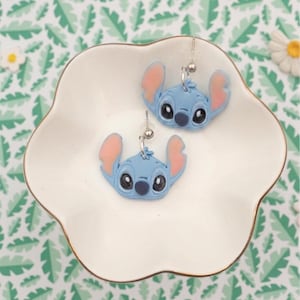 Pu&ograve; includere: Un paio di orecchini blu di Stitch con orecchie rosa e occhi neri, appoggiati su un piatto bianco con bordo smerlato. Gli orecchini sono argentati e pendono da una piccola sfera. Lo sfondo presenta un motivo a foglie verdi e bianche.