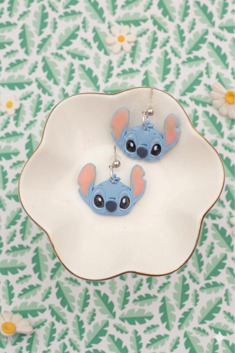 Pu&ograve; includere: Un paio di orecchini blu di Stitch con orecchie rosa e occhi neri, appoggiati su un piatto bianco con bordo smerlato. Gli orecchini sono argentati e pendono da una piccola sfera. Lo sfondo presenta un motivo a foglie verdi e bianche.