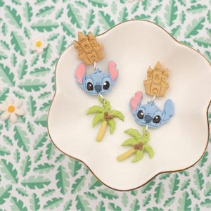 May include: A pair of blue Stitch earrings with pink ears and black eyes, each with a tan castle charm and a green palm tree charm. The earrings are displayed on a white, scalloped dish with a gold rim, set against a green and white leaf pattern.