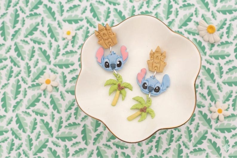 May include: A pair of blue Stitch earrings with pink ears and black eyes, each with a tan castle charm and a green palm tree charm. The earrings are displayed on a white, scalloped dish with a gold rim, set against a green and white leaf pattern.