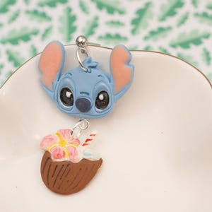 May include: A pair of dangle earrings featuring a blue cartoon character head with large black eyes and pink ears, and a brown coconut drink with a pink flower and a red and white straw. The earrings are on a white plate.