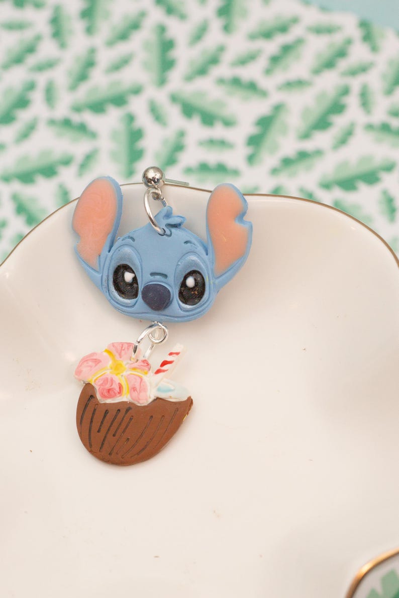 May include: A pair of dangle earrings featuring a blue cartoon character head with large black eyes and pink ears, and a brown coconut drink with a pink flower and a red and white straw. The earrings are on a white plate.