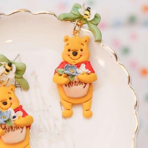 May include: Yellow Winnie the Pooh earrings with red shirts, holding blue and white flowers and a honey pot that says "Hunny". Each earring has a green bow with a bee attached.