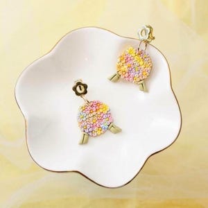 May include: A pair of floral earrings in pastel shades, displayed on a white, scalloped dish. The earrings have small, colourful flowers and gold-toned details. The background is a soft yellow.