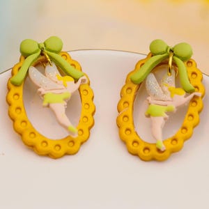 May include: A pair of whimsical earrings featuring a fairy design. Each earring has a yellow oval frame with a scalloped edge, a green bow, and a fairy with glittery wings and a light green dress. The earrings are on a white surface.