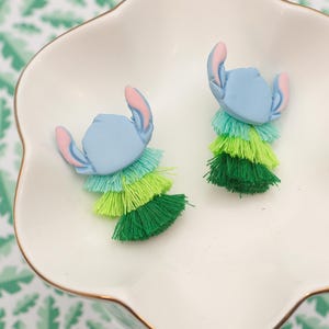 May include: Two blue character-shaped earrings with pink ears and layered green tassels, presented in a white, scalloped dish with a gold rim. The background features a green and white leaf pattern. These earrings offer a playful, colourful design.