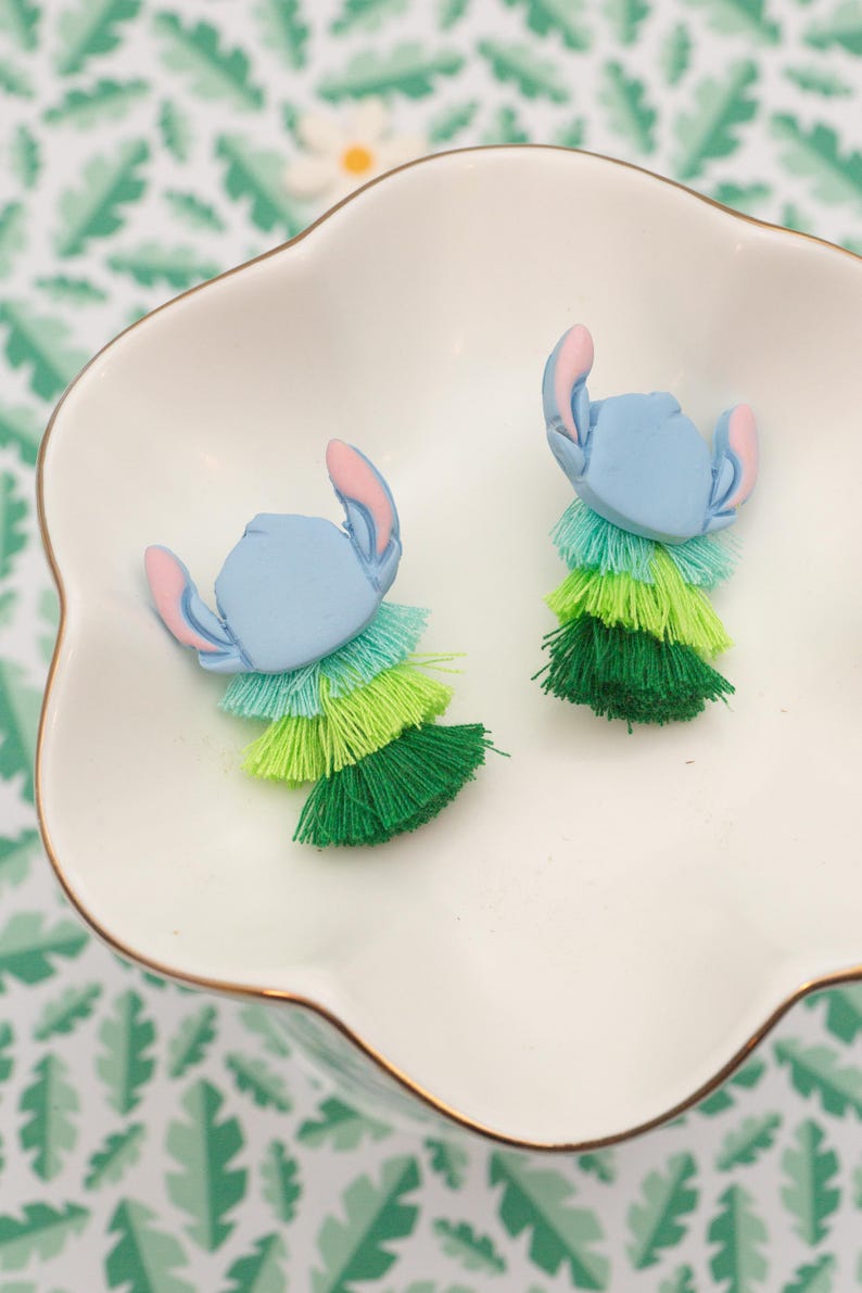 May include: Two blue character-shaped earrings with pink ears and layered green tassels, presented in a white, scalloped dish with a gold rim. The background features a green and white leaf pattern. These earrings offer a playful, colourful design.