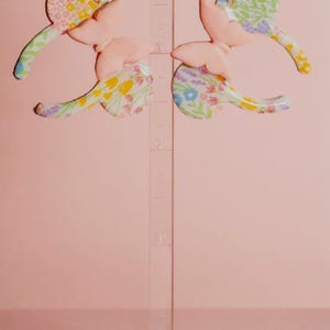 K&ouml;nnte beinhalten: Ein Paar pastellrosa Schmetterlingsohrringe mit floralen Akzenten. Die Ohrringe werden auf einem transparenten St&auml;nder vor einem zartrosa Hintergrund pr&auml;sentiert. Die Schmetterlingsfl&uuml;gel haben ein zartes, farbenfrohes Blumenmuster.
