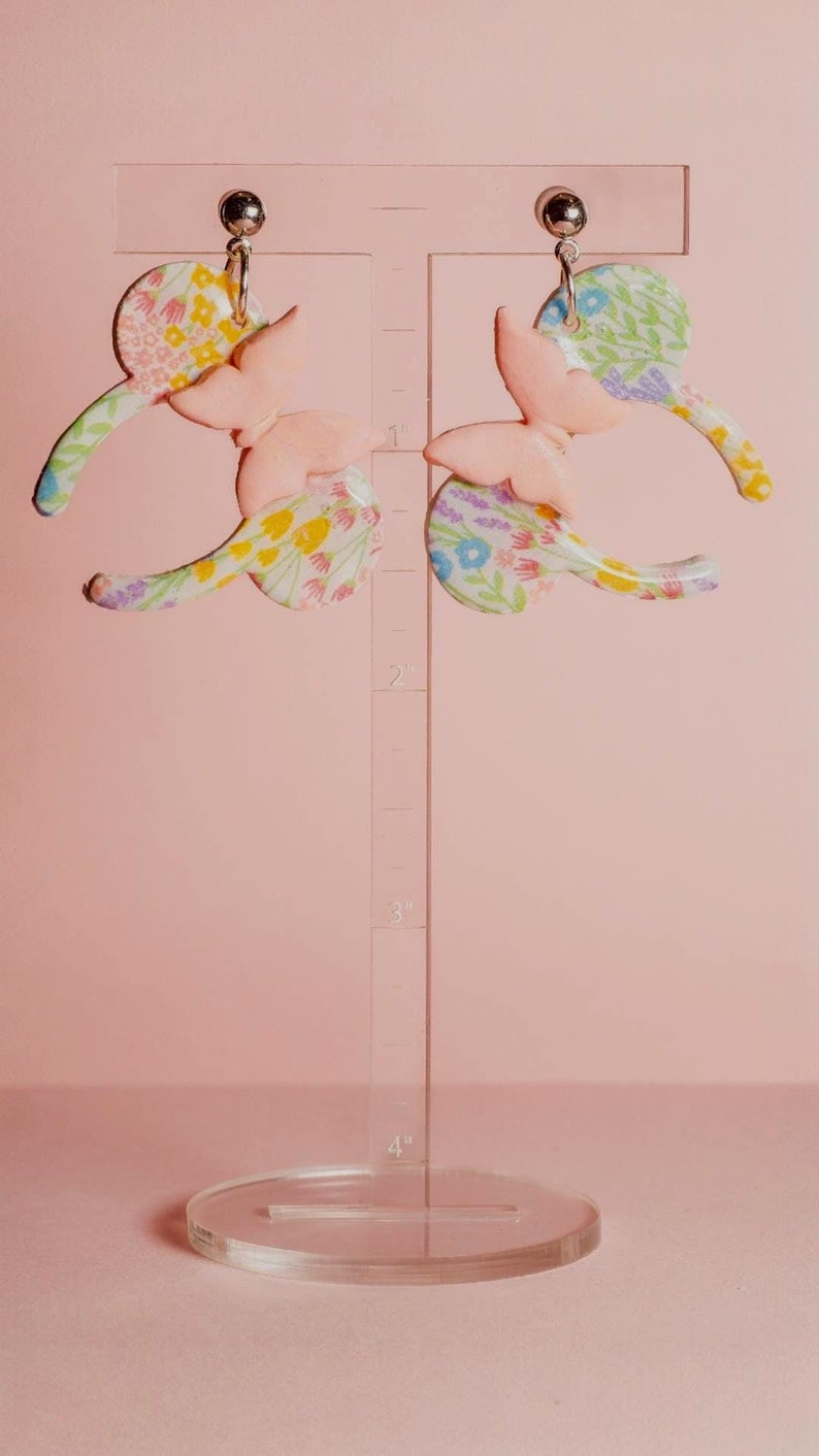 K&ouml;nnte beinhalten: Ein Paar pastellrosa Schmetterlingsohrringe mit floralen Akzenten. Die Ohrringe werden auf einem transparenten St&auml;nder vor einem zartrosa Hintergrund pr&auml;sentiert. Die Schmetterlingsfl&uuml;gel haben ein zartes, farbenfrohes Blumenmuster.