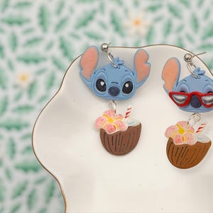 May include: Two pairs of novelty earrings featuring a blue cartoon character with large ears. One pair has the character without glasses, and the other has red glasses. Each earring has a brown coconut with a pink flower and a red and white straw.