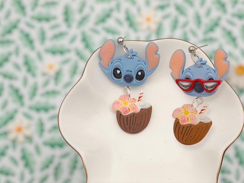 May include: Two pairs of novelty earrings featuring a blue cartoon character with large ears. One pair has the character without glasses, and the other has red glasses. Each earring has a brown coconut with a pink flower and a red and white straw.