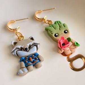 May include: A pair of gold hoop earrings with charm pendants. One earring features a raccoon character in blue and orange clothing. The other earring has a Groot character holding a pink flower. The earrings are on a white surface.