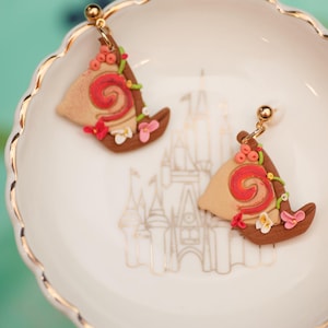 May include: A pair of handmade earrings featuring a tan sail design with a red spiral, brown accents, and floral details. The earrings are displayed on a white scalloped dish with a gold rim.