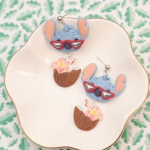 May include: A collection of handmade earrings featuring a blue alien character wearing red sunglasses and coconut drink earrings with pink flowers. The earrings are displayed on a white plate with a gold rim, set against a green leaf pattern.