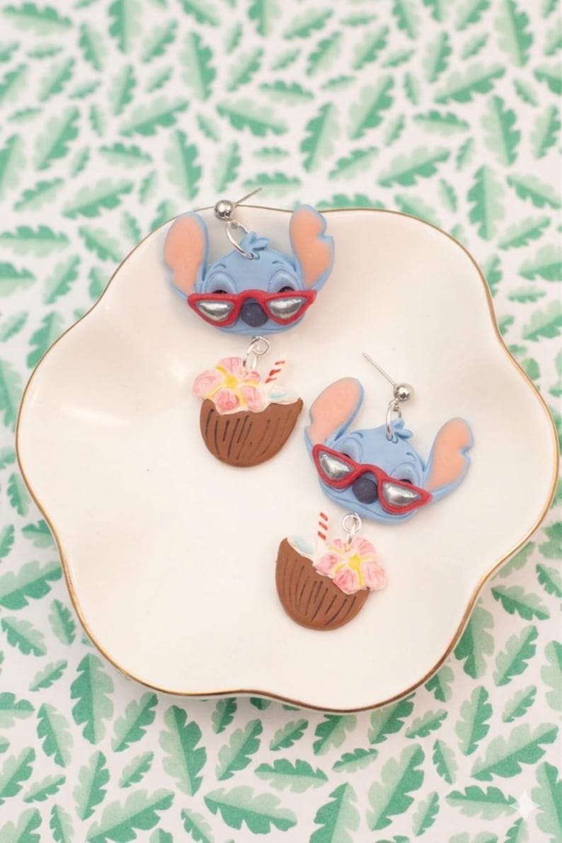 May include: A collection of handmade earrings featuring a blue alien character wearing red sunglasses and coconut drink earrings with pink flowers. The earrings are displayed on a white plate with a gold rim, set against a green leaf pattern.