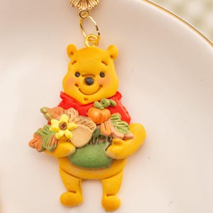 May include: A gold-toned earring featuring a yellow bear character wearing a red shirt, holding a green pot filled with autumn-themed items including an acorn, a small orange pumpkin, and a yellow flower, suspended from a gold daisy stud.