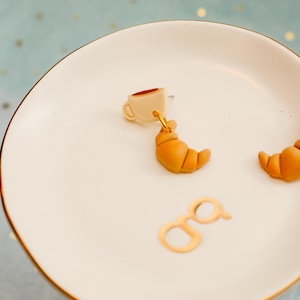 May include: A pair of novelty earrings featuring a coffee cup and croissant design. The earrings are gold-toned and dangle from a small beige coffee cup with a brown interior. A golden-brown croissant hangs below the cup. The earrings rest on a white plate.