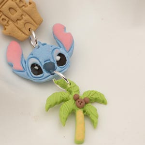 May include: A blue character head with pink ears and black eyes is connected to a green palm tree charm with a brown mouse. A tan castle charm is also attached. The charms are on a white surface.