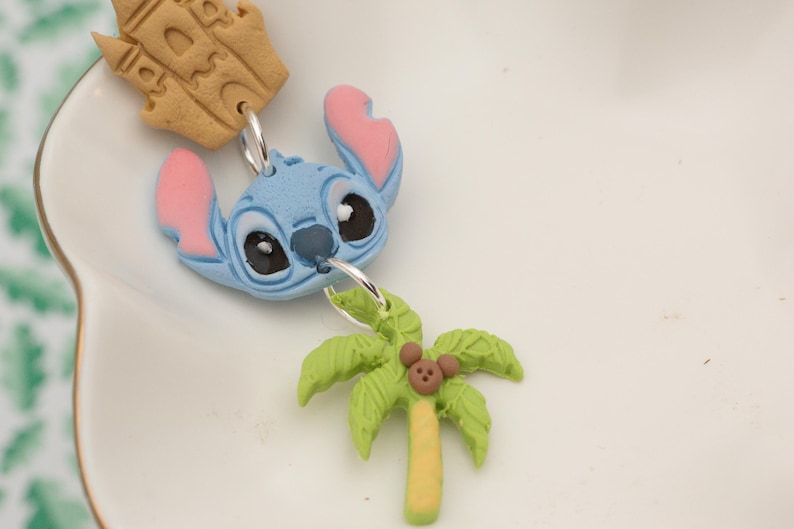 May include: A blue character head with pink ears and black eyes is connected to a green palm tree charm with a brown mouse. A tan castle charm is also attached. The charms are on a white surface.