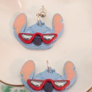 Pu&ograve; includere: Un paio di orecchini blu di Stitch. Ogni orecchino presenta un personaggio di Stitch blu chiaro con orecchie rosa e un paio di occhiali da sole con montatura rossa e lenti argentate. Gli orecchini sono esposti su un piatto bianco con bordo dorato.