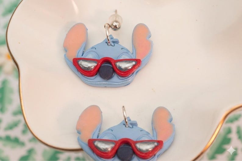 Pu&ograve; includere: Un paio di orecchini blu di Stitch. Ogni orecchino presenta un personaggio di Stitch blu chiaro con orecchie rosa e un paio di occhiali da sole con montatura rossa e lenti argentate. Gli orecchini sono esposti su un piatto bianco con bordo dorato.