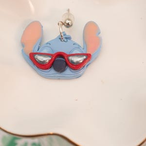 Pu&ograve; includere: Un orecchino Stitch azzurro con orecchie rosa e occhiali da sole rossi. L'orecchino &egrave; un design di personaggi dei cartoni animati. L'orecchino &egrave; su un piatto bianco smerlato con bordo dorato.