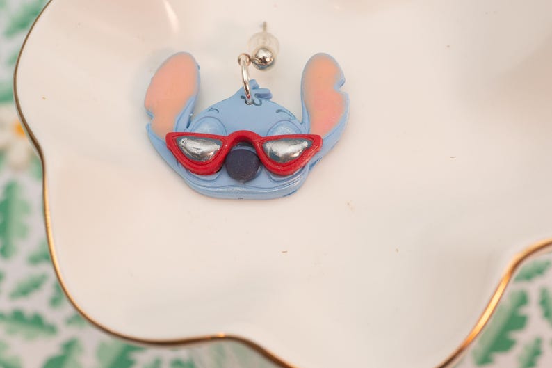 Pu&ograve; includere: Un orecchino Stitch azzurro con orecchie rosa e occhiali da sole rossi. L'orecchino &egrave; un design di personaggi dei cartoni animati. L'orecchino &egrave; su un piatto bianco smerlato con bordo dorato.