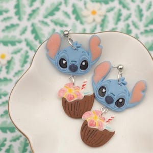 May include: A pair of blue Stitch earrings with pink ears and a coconut drink charm. The earrings are on a white, scalloped dish against a green and white floral background.