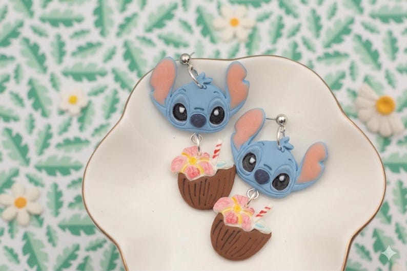May include: A pair of blue Stitch earrings with pink ears and a coconut drink charm. The earrings are on a white, scalloped dish against a green and white floral background.