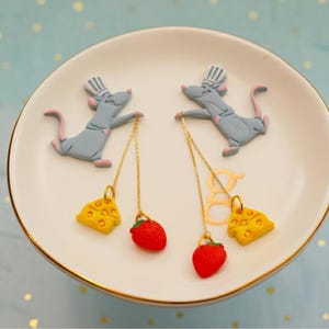 May include: Novelty earrings with a cartoon rat chef design. Each earring has a gold-coloured chain suspending a yellow cheese wedge and a red strawberry charm. The earrings are displayed on a white plate with a gold trim.