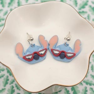 Pu&ograve; includere: Un paio di orecchini blu a forma di personaggio con orecchie rosa e occhiali da sole rossi, appoggiati in un piatto bianco smerlato. Gli orecchini hanno perni in metallo color argento. Lo sfondo presenta un motivo a foglie verdi e bianche.