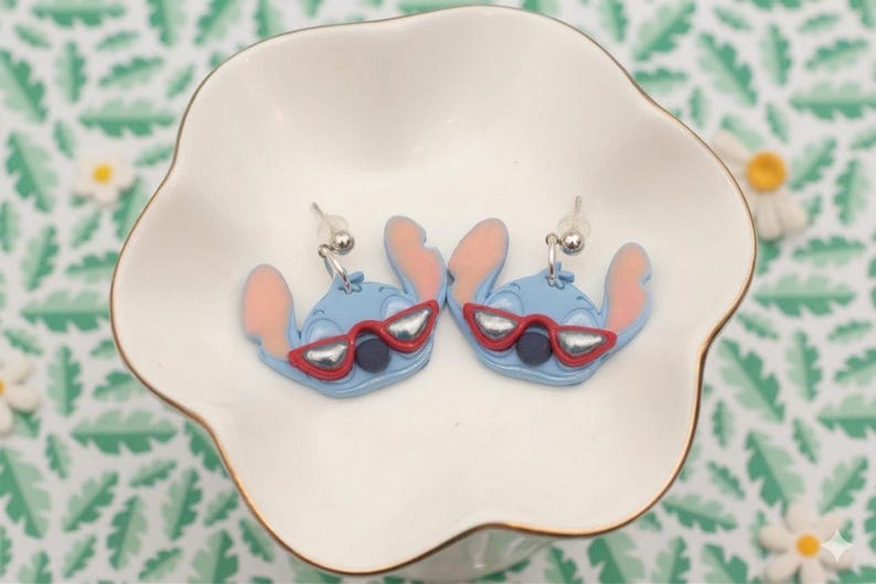 Pu&ograve; includere: Un paio di orecchini blu a forma di personaggio con orecchie rosa e occhiali da sole rossi, appoggiati in un piatto bianco smerlato. Gli orecchini hanno perni in metallo color argento. Lo sfondo presenta un motivo a foglie verdi e bianche.