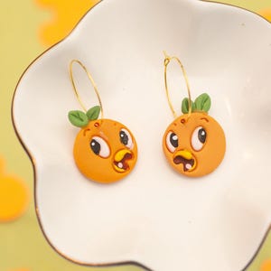 May include: A pair of orange-shaped earrings with cartoon faces, green leaf accents, and gold-colored hoops. The earrings are displayed on a white, scalloped-edge dish against a yellow background with orange shapes.