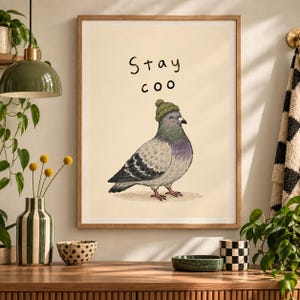 Puede incluir: Una lámina enmarcada que muestra una paloma gris con un pequeño gorro de punto verde. La ilustración está sobre un fondo crema claro con el texto Stay coo escrito en letras negras dibujadas a mano sobre el ave.