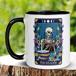 May include: White ceramic coffee mug with a black rim and handle. The mug features a tarot card design with a skeleton reading a book. The card is titled "The Reader" and has a blue background with stars and moons. The skeleton is surrounded by flowers.