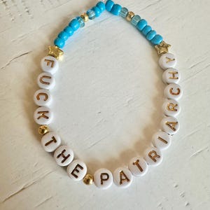 May include: A white beaded bracelet with gold lettering spelling "F**K THE PATRIARCHY". The bracelet has blue beads on one side and white beads on the other.