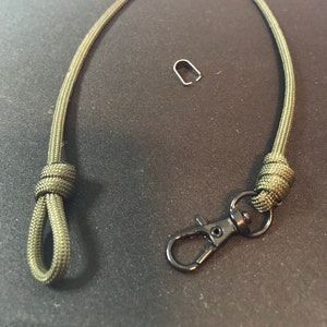 May include: A green paracord lanyard with a black metal clip and a small metal loop. The lanyard is knotted at both ends.