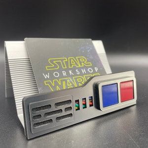 May include: A black and silver business card holder with the text "STAR WORKSHOP WARS". The holder has a rectangular design with a blue and red square on the front. The holder is on a black surface.