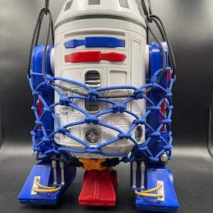 May include: A white and blue R2-D2-shaped cooler with a black and blue mesh net and a black strap. The cooler has red and blue details on the front and yellow accents on the bottom.