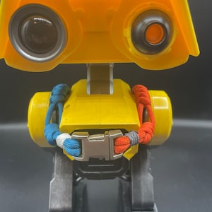 May include: A yellow robot toy with large, round eye-like features and a rectangular head. It has a silver buckle and braided rope details in blue, white, and red. The robot's legs are dark gray.